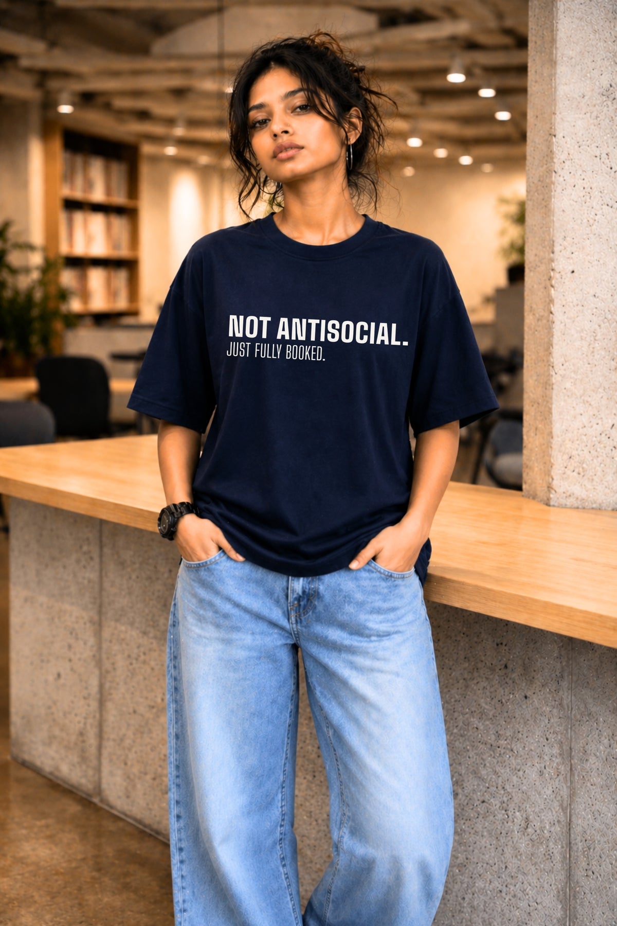 Woman wearing navy oversized graphic tee "Not Antisocial. Just Fully Booked." in a library setting — JUNOSHI Unfiltered Collection
