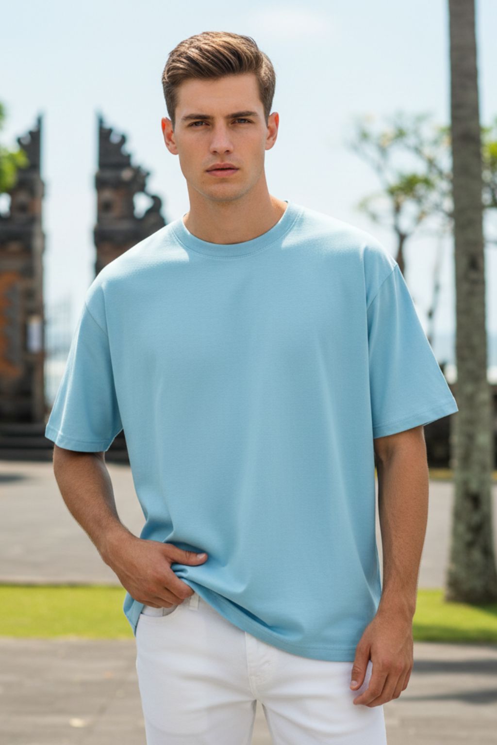 Man wearing a baby blue oversized t-shirt and white pants outdoors with trees and a clear sky in the background