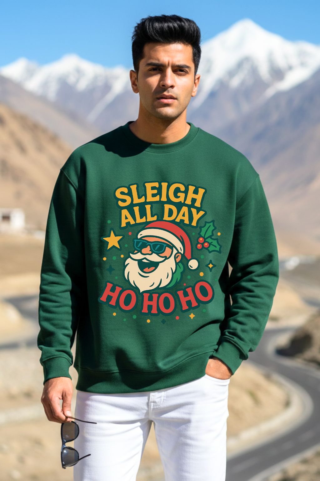 Man wearing a bottle green oversized sweatshirt with Christmas-themed text and graphics, standing in a mountainous landscape.