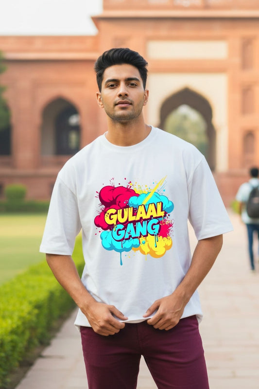 Man wearing a white oversized t-shirt with colorful text 'Gulaal Gang' in front of a brick building.