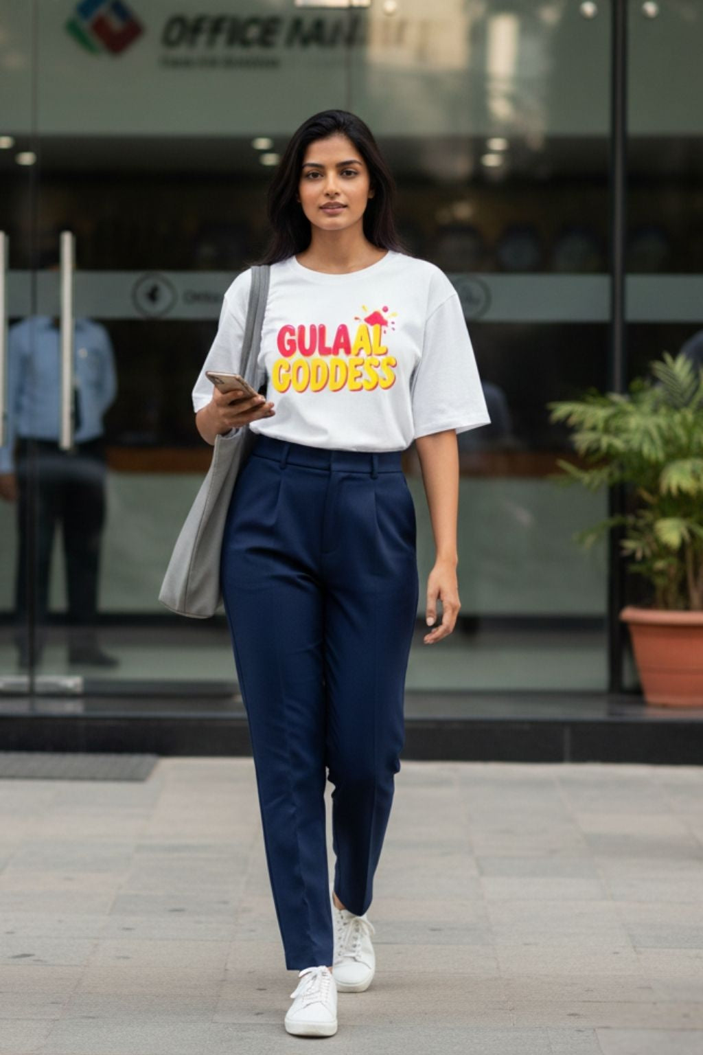 lifestyle shot view of a woman wearing a white oversized t-shirt with 'Gulaal Goddess' text, navy pants, and white sneakers walking outdoors.