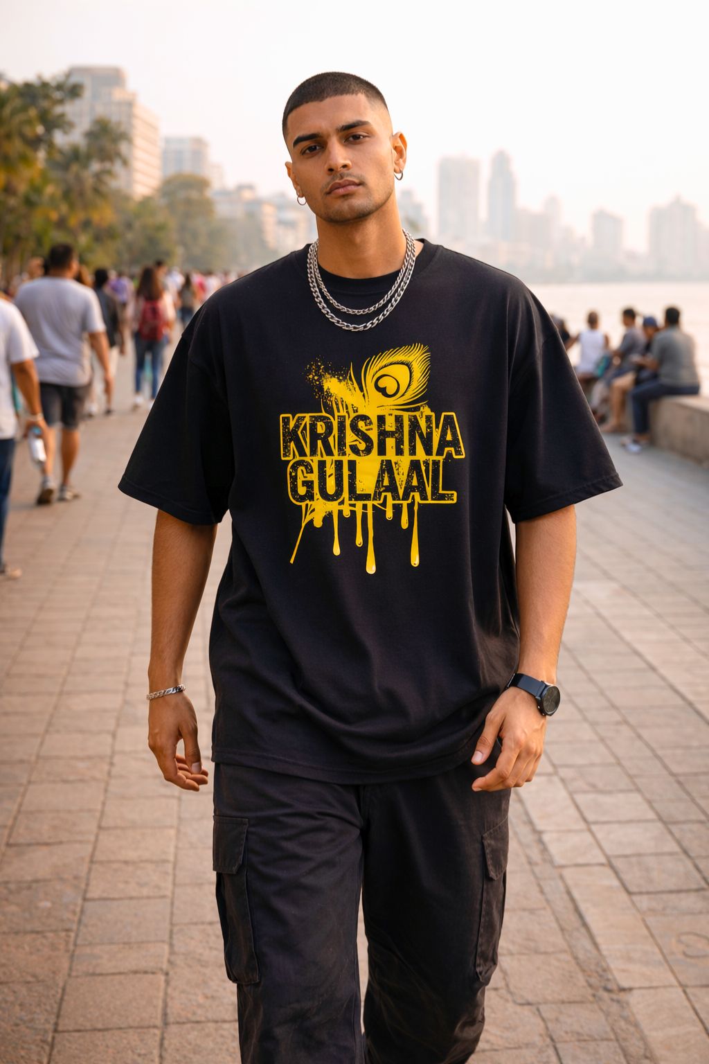 Man wearing a black oversized t-shirt with yellow text "Krishna Gulaal" walking on a city street.