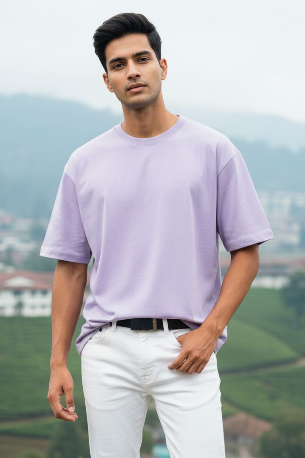 Man wearing a lavendar oversized  t-shirt and white pants standing outdoors with mountains in the background