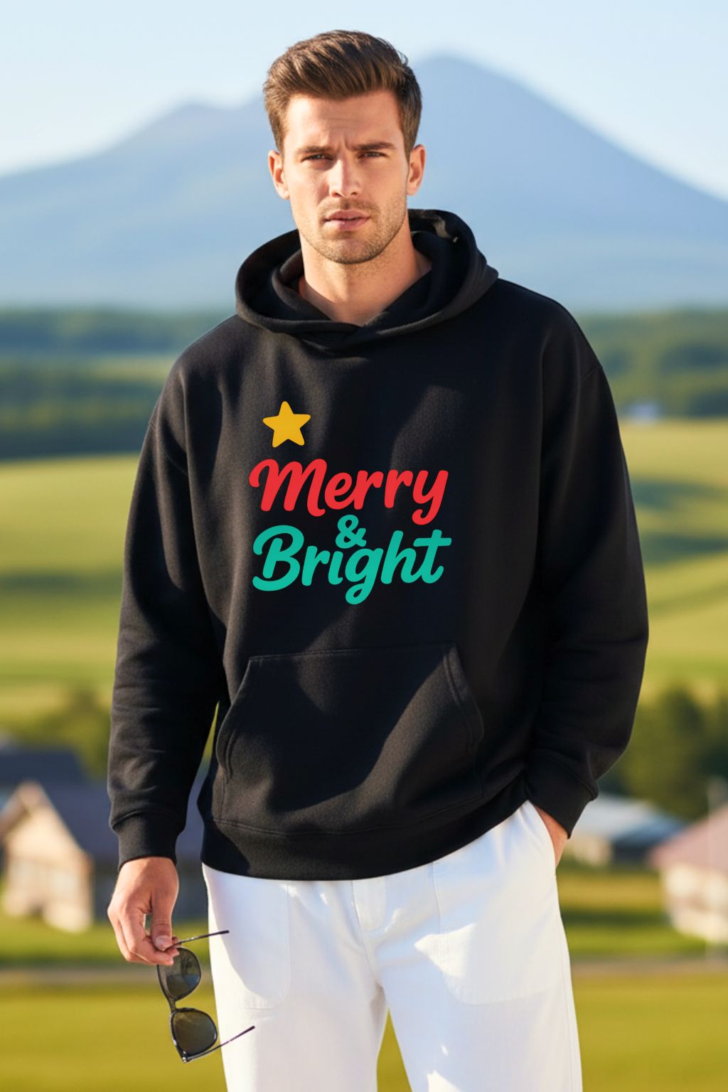 Man wearing a black oversized hoodie with 'Merry & Bright' text, standing outdoors with greenery and mountains in the background.