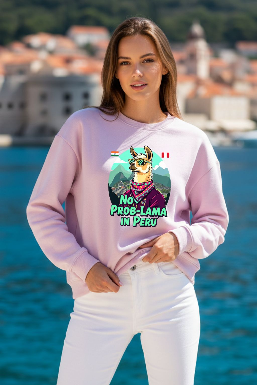 Woman wearing a light baby pink oversized sweatshirt with a llama graphic and text, standing by a waterfront with buildings in the background.