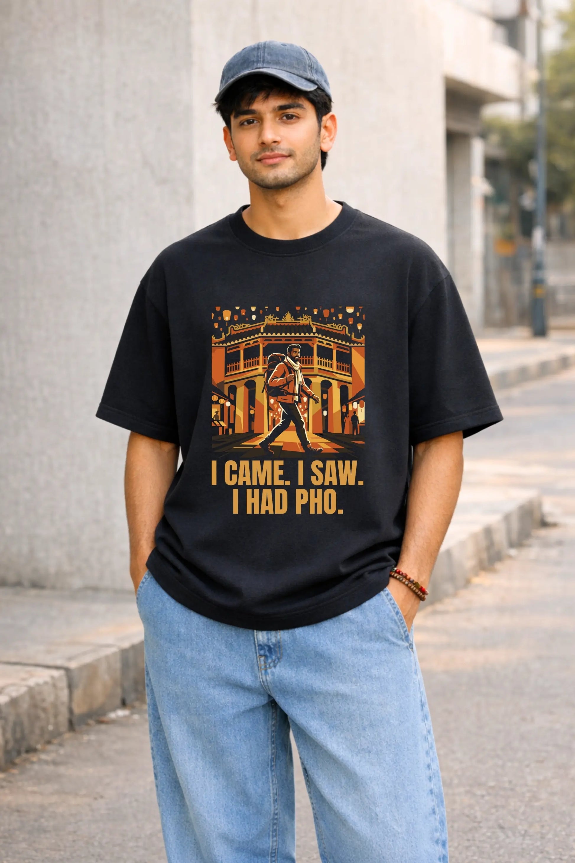 Indian man wearing black oversized graphic tee with Hội An Vietnam lantern street print and I Came I Saw I Had Pho bold text, styled with blue jeans and cap, JUNOSHI