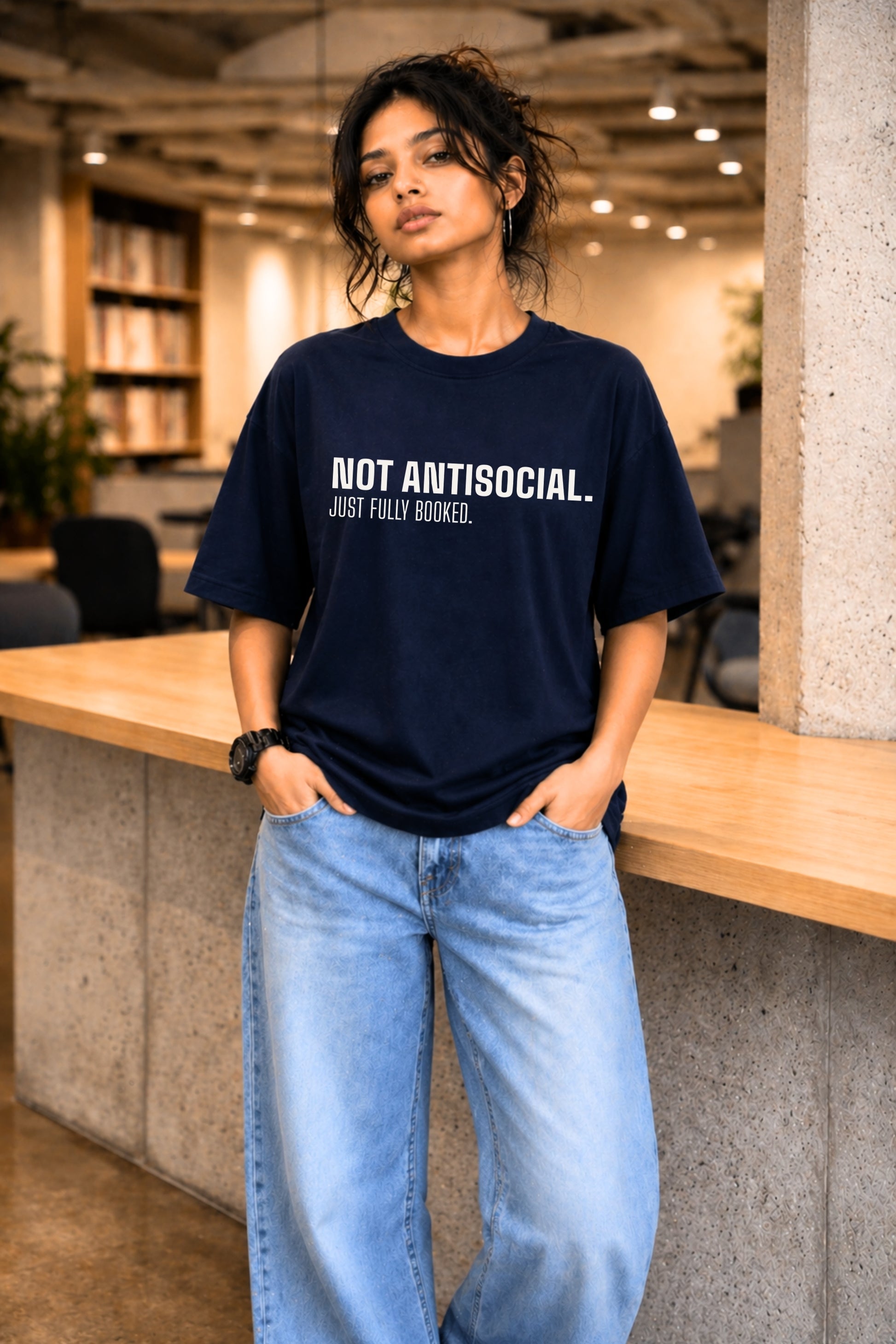 Woman wearing navy oversized graphic tee "Not Antisocial. Just Fully Booked." in a library setting — JUNOSHI Unfiltered Collection