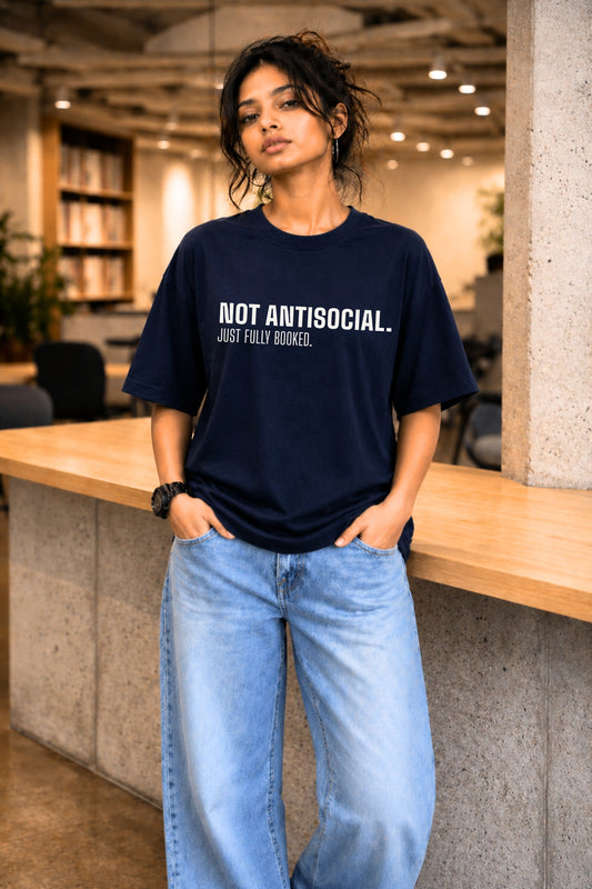 Woman wearing navy oversized graphic tee "Not Antisocial. Just Fully Booked." in a library setting — JUNOSHI Unfiltered Collection