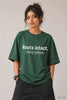 Woman wearing JUNOSHI Roots Intact Ceiling Removed oversized tee in forest green — bold white typography, relaxed drop-shoulder fit, styled with black cargo pants