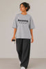 Becoming. Do Not Narrate oversized tee in grey — women's full body front view, styled with wide-leg black pants | JUNOSHI Unfiltered Collection