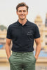 Man wearing JUNOSHI Made of Stars black men's polo with constellation graphic and 
'Made of Stars' text on chest, styled with olive green trousers, against a blurred 
Indian heritage architecture background.
