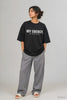 Woman wearing black oversized tee with bold white text reading My Energy My Intake Policy, styled with grey wide-leg pants and white slides, part of JUNOSHI Unfiltered drop